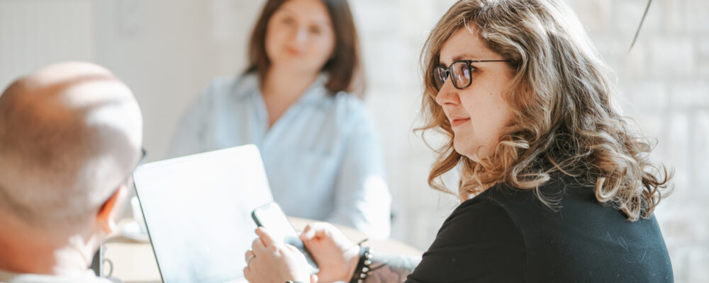 Frau mit Brille haelt Smartphone, Laptop auf dem Tisch.
